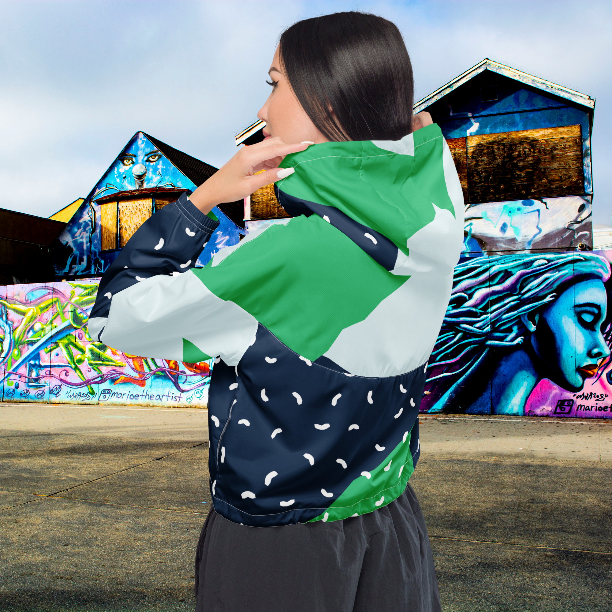 Ein Damen Cropped Windbreaker mit einem modernen, geometrischen Blockmuster in Dunkelblau, hellem Mintgrün und strahlendem Grün. Der dunkelblaue Bereich ist zusätzlich mit kleinen weißen, unregelmäßigen Strichformen versehen. Die Jacke hat eine Kapuze, einen halben Reißverschluss mit weißen Kordeln und einen verstellbaren Saum. Mockup Girl Rücken Ansicht. By Just a Girl.