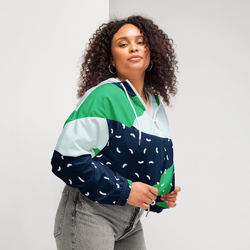 Ein Damen Cropped Windbreaker mit einem modernen, geometrischen Blockmuster in Dunkelblau, hellem Mintgrün und strahlendem Grün. Der dunkelblaue Bereich ist zusätzlich mit kleinen weißen, unregelmäßigen Strichformen versehen. Die Jacke hat eine Kapuze, einen halben Reißverschluss mit weißen Kordeln und einen verstellbaren Saum. Mockup woman seitliche Ansicht rechts. By Just a Girl.