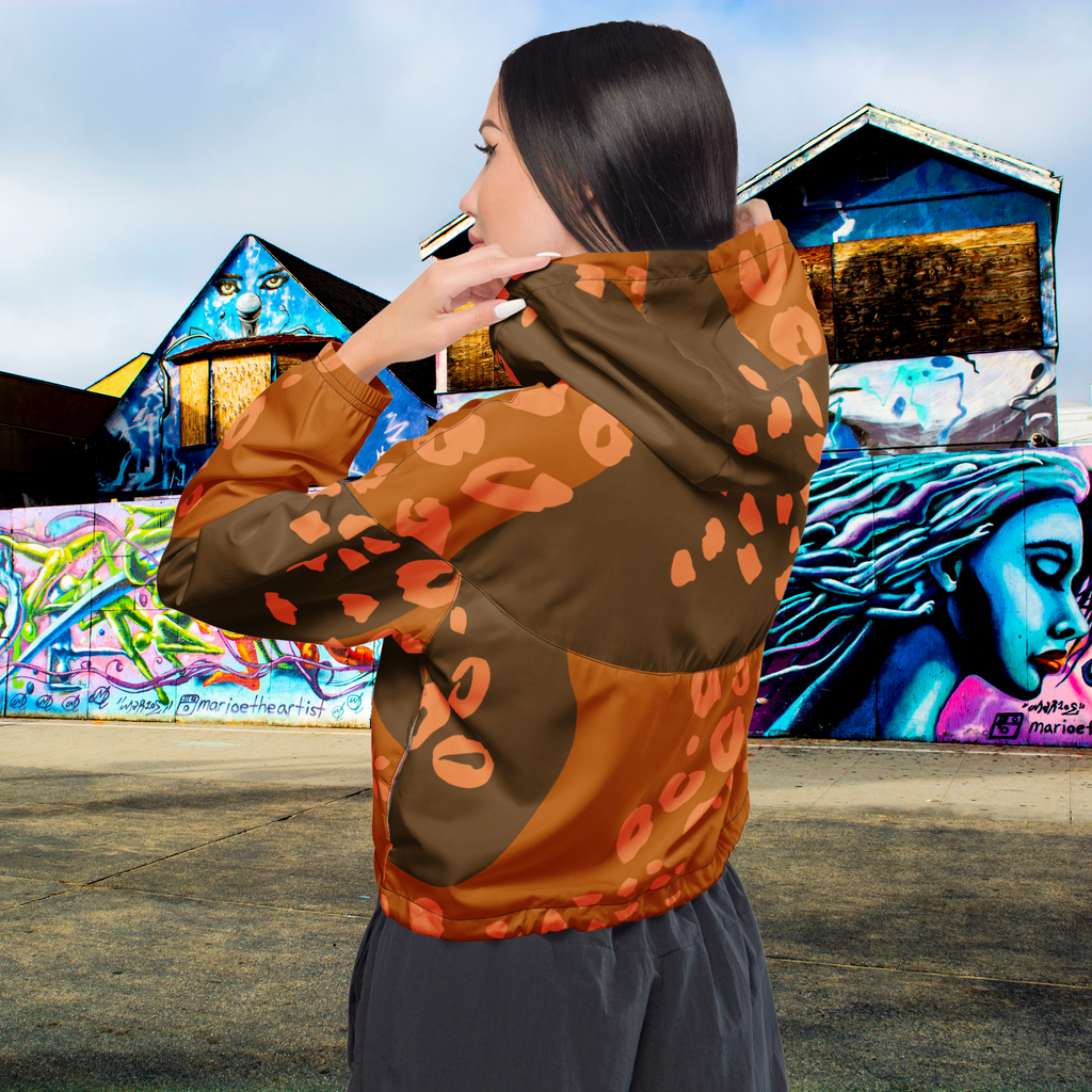 Ein Damen Cropped Windbreaker in Dunkelbraun und Orange mit einem abstrakten Muster aus unregelmäßigen Flecken und Formen, teilweise leopardenähnlich. Die Jacke hat eine Kapuze, einen halben Reißverschluss, schwarze Kordeln und einen verstellbaren Saum. Mockup Girl Rückenansicht. By Just a Girl.