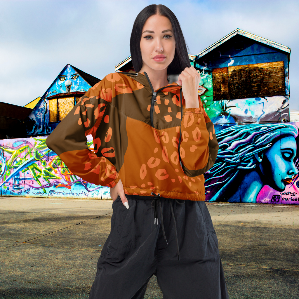 Ein Damen Cropped Windbreaker mit schwarzen Reissverschluss  in Dunkelbraun und Orange mit einem abstrakten Muster aus unregelmäßigen Flecken und Formen, teilweise leopardenähnlich. Die Jacke hat eine Kapuze, einen halben Reißverschluss, schwarze Kordeln und einen verstellbaren Saum. Mockup Girl Vorderansicht. By Just a Girl.