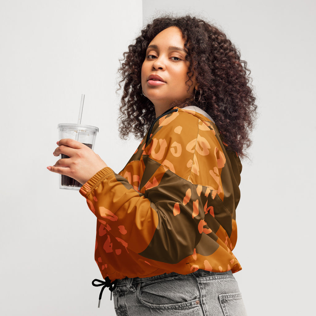 Ein Damen Cropped Windbreaker in Dunkelbraun und Orange mit einem abstrakten Muster aus unregelmäßigen Flecken und Formen, teilweise leopardenähnlich. Die Jacke hat eine Kapuze, einen halben Reißverschluss, schwarze Kordeln und einen verstellbaren Saum. Mockup woman seitliche Ansicht links. By Just a Girl.