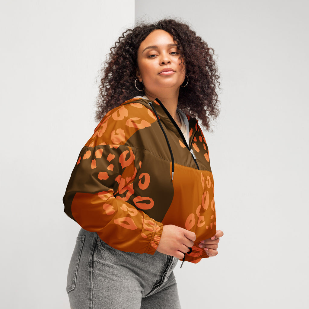 Ein Damen Cropped Windbreaker in Dunkelbraun und Orange mit einem abstrakten Muster aus unregelmäßigen Flecken und Formen, teilweise leopardenähnlich. Die Jacke hat eine Kapuze, einen halben Reißverschluss, schwarze Kordeln und einen verstellbaren Saum. Mockup woman seitliche Ansicht rechts. By Just a Girl.