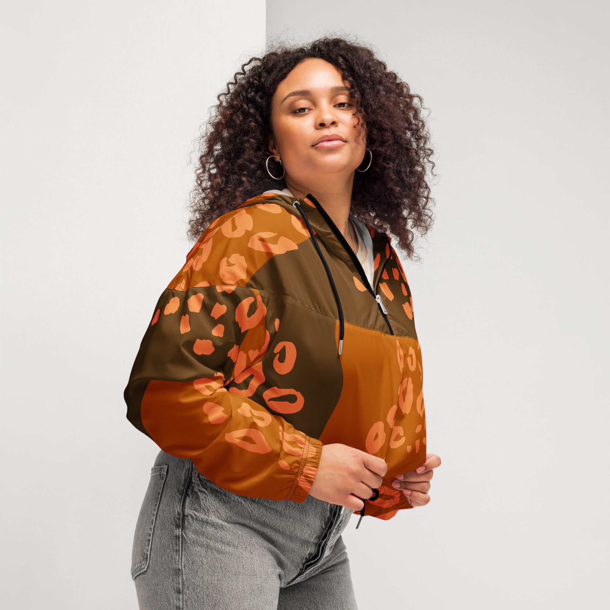 Ein Damen Cropped Windbreaker in Dunkelbraun und Orange mit einem abstrakten Muster aus unregelmäßigen Flecken und Formen, teilweise leopardenähnlich. Die Jacke hat eine Kapuze, einen halben Reißverschluss, schwarze Kordeln und einen verstellbaren Saum. Mockup woman seitliche Ansicht rechts. By Just a Girl.