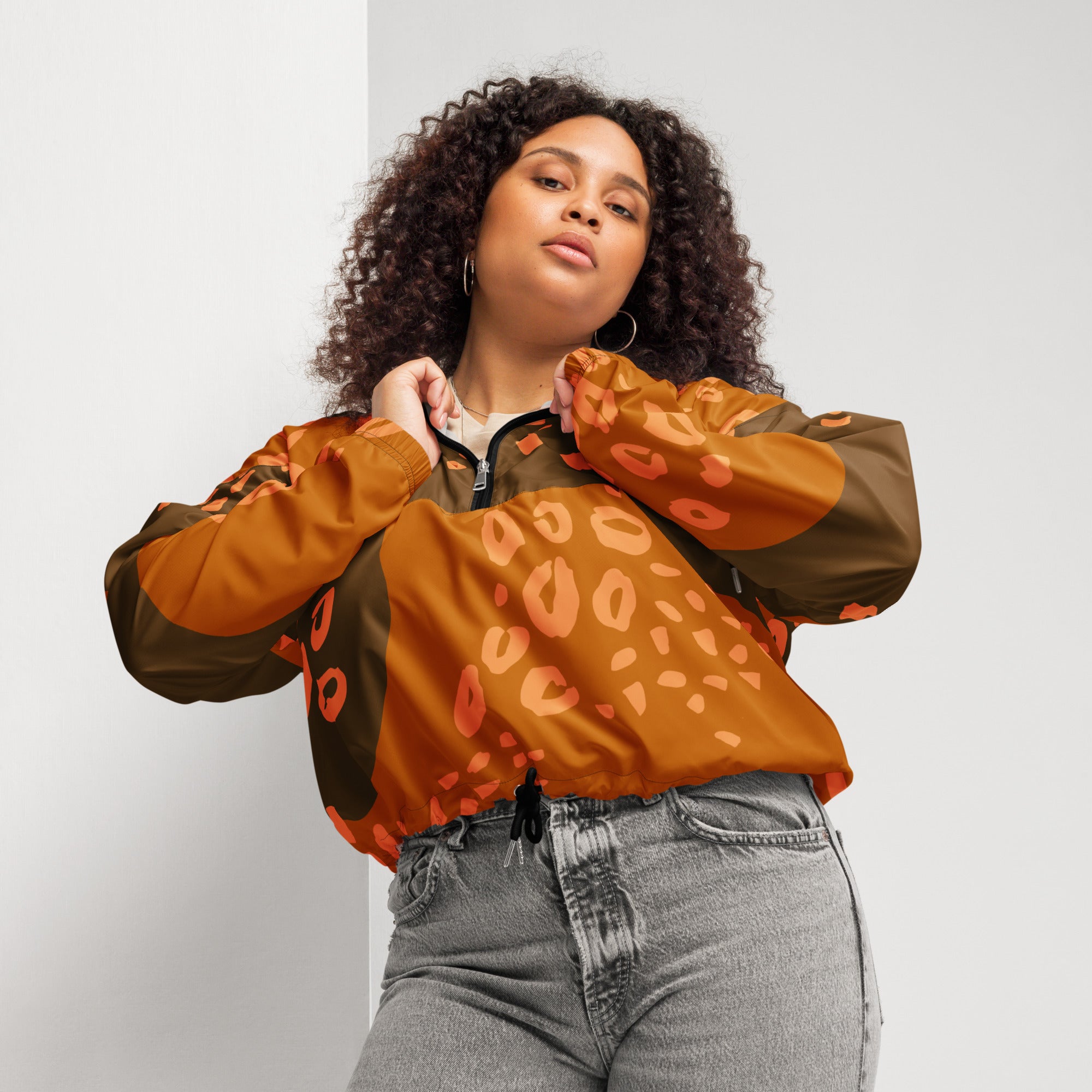 Ein Damen Cropped Windbreaker in Dunkelbraun und Orange mit einem abstrakten Muster aus unregelmäßigen Flecken und Formen, teilweise leopardenähnlich. Die Jacke hat eine Kapuze, einen halben Reißverschluss, schwarze Kordeln und einen verstellbaren Saum. Mockup Woman Vorderansicht. By Just a Girl.