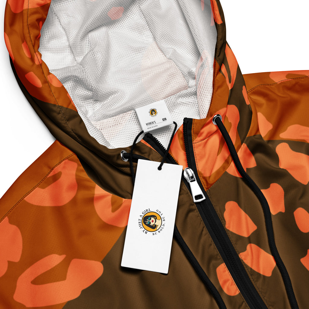 Ein Damen Cropped Windbreaker in Dunkelbraun und Orange mit einem abstrakten Muster aus unregelmäßigen Flecken und Formen, teilweise leopardenähnlich. Die Jacke hat eine Kapuze, einen halben Reißverschluss, schwarze Kordeln und einen verstellbaren Saum. Mockup Kapuzen Ansicht mit schwarzem Reißverschluß. By Just a Girl