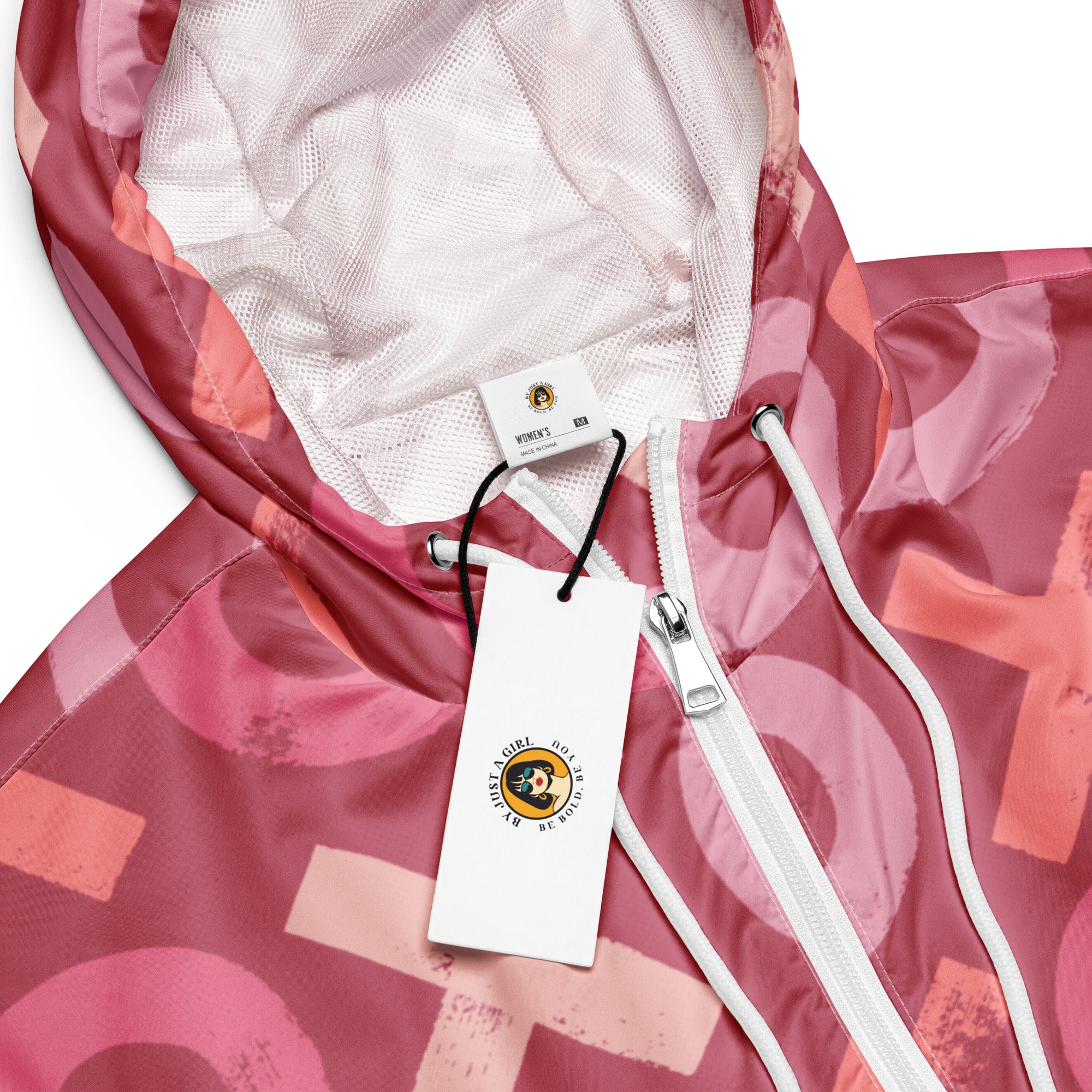 Ein Damen Cropped Windbreaker in verschiedenen Rosa- und Rottönen mit einem durchgängigen Muster aus großen X- und O-Formen. Die Jacke hat eine Kapuze, einen halben Reißverschluss, weiße Kordeln und einen verstellbaren Saum. by Just a Girl.