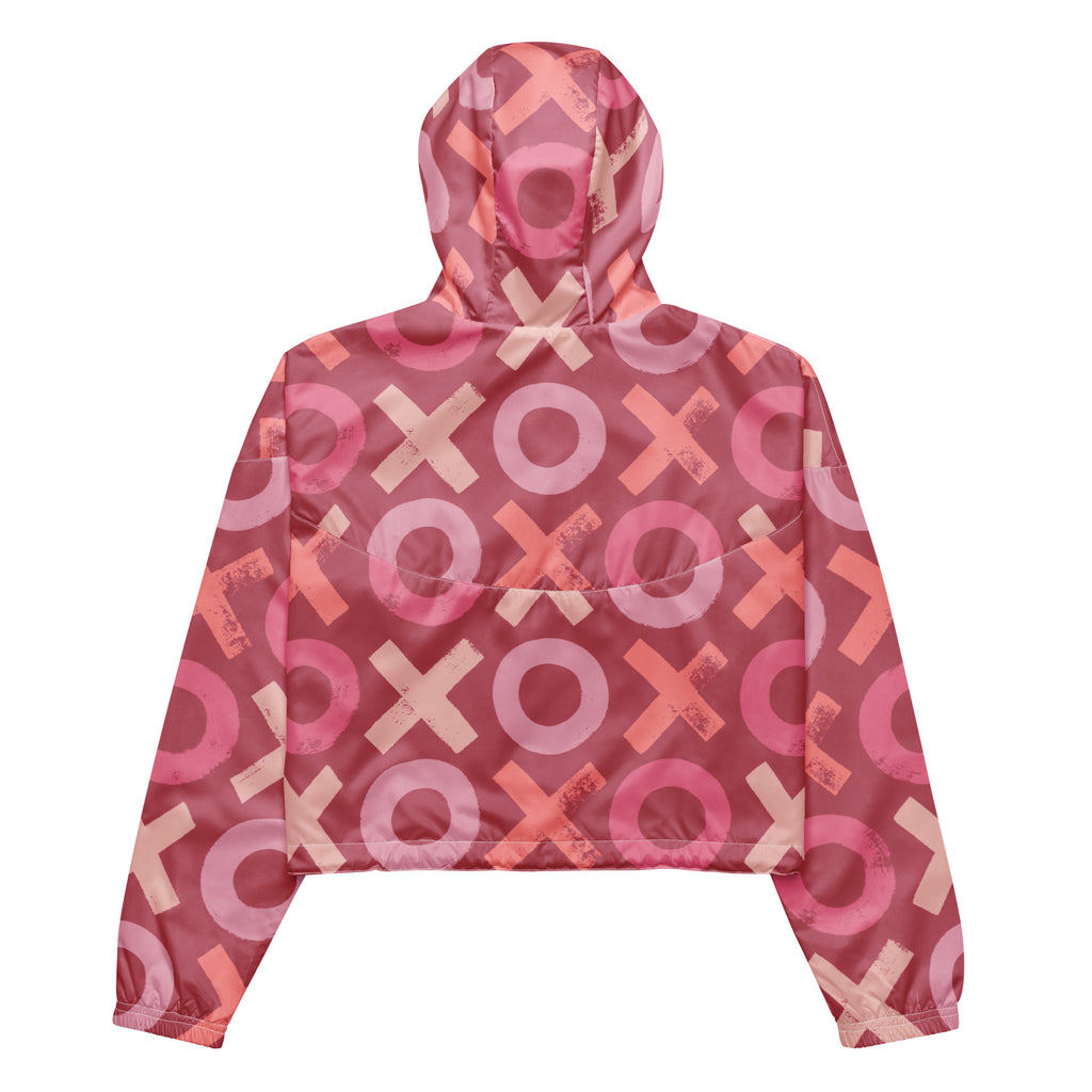 Ein Damen Cropped Windbreaker in verschiedenen Rosa- und Rottönen mit einem durchgängigen Muster aus großen X- und O-Formen. Die Jacke hat eine Kapuze, einen halben Reißverschluss, weiße Kordeln und einen verstellbaren Saum. Die Jacke ist flach ausgebreitet. Präsentiert von einer Frau in der Frontansicht by Just a Girl.