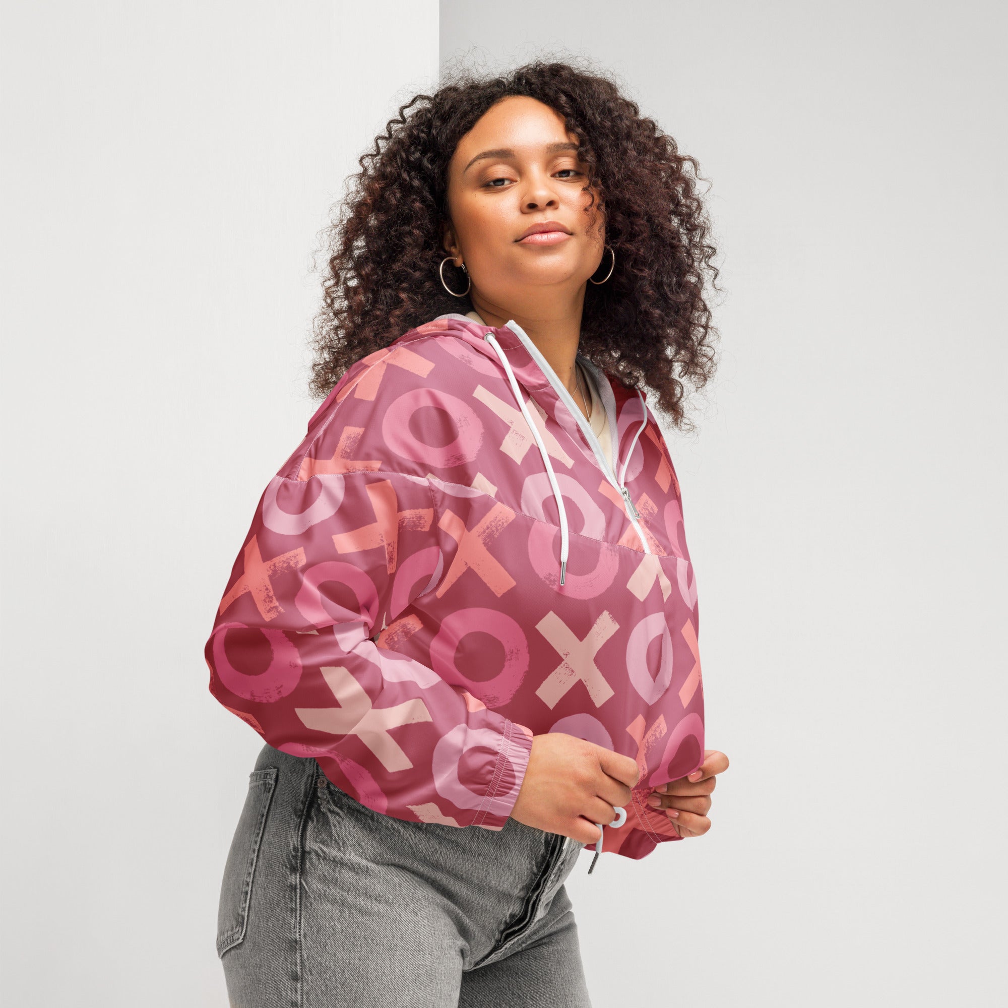 Ein Damen Cropped Windbreaker in verschiedenen Rosa- und Rottönen mit einem durchgängigen Muster aus großen X- und O-Formen. Die Jacke hat eine Kapuze, einen halben Reißverschluss, weiße Kordeln und einen verstellbaren Saum. Die Jacke ist flach ausgebreitet. Präsentiert von einer kräftigeren Frau in der Seitenansicht by Just a Girl.