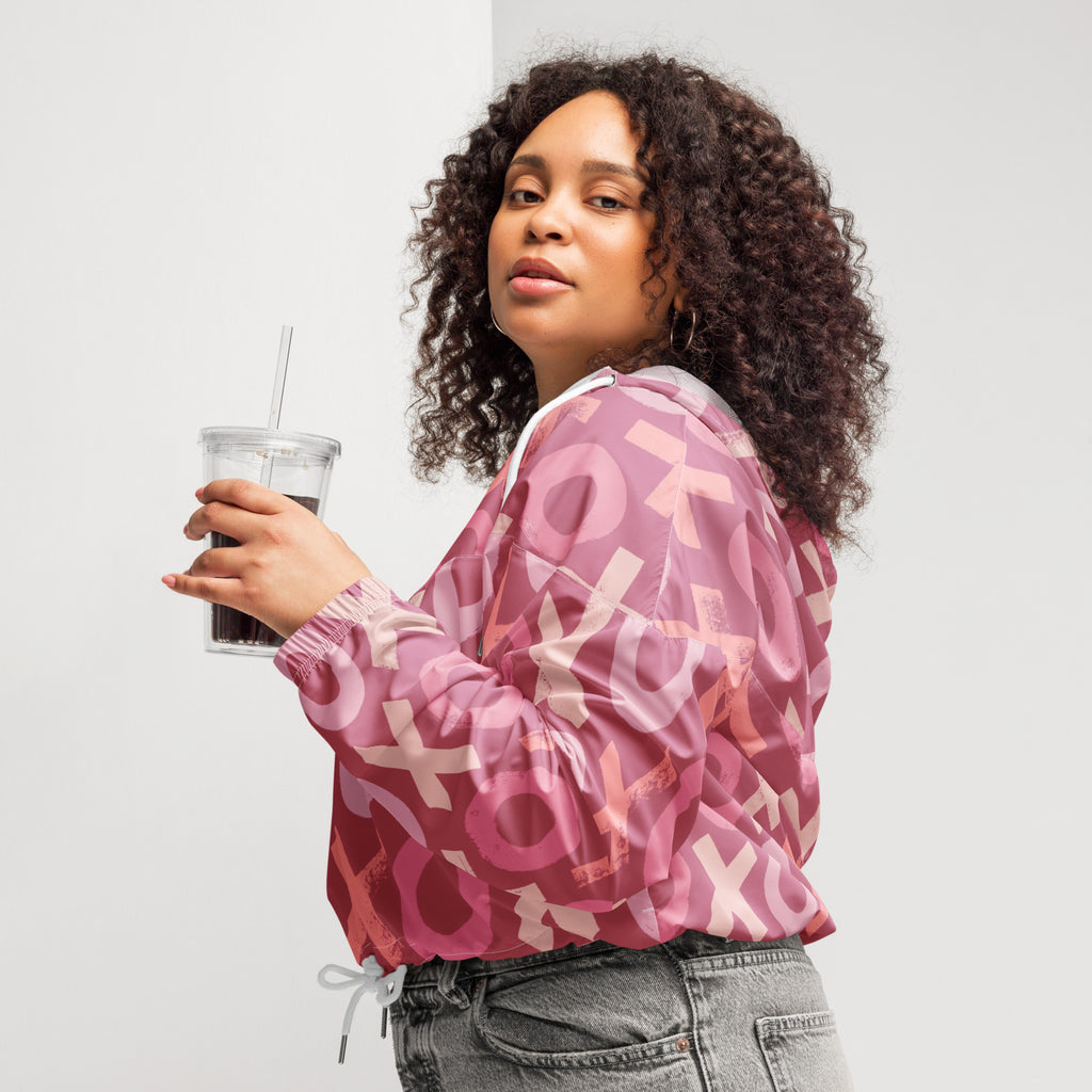 Ein Damen Cropped Windbreaker in verschiedenen Rosa- und Rottönen mit einem durchgängigen Muster aus großen X- und O-Formen. Die Jacke hat eine Kapuze, einen halben Reißverschluss, weiße Kordeln und einen verstellbaren Saum. Die Jacke ist flach ausgebreitet. Präsentiert von einer Frau in der seitlichen Ansicht by Just a Girl.
