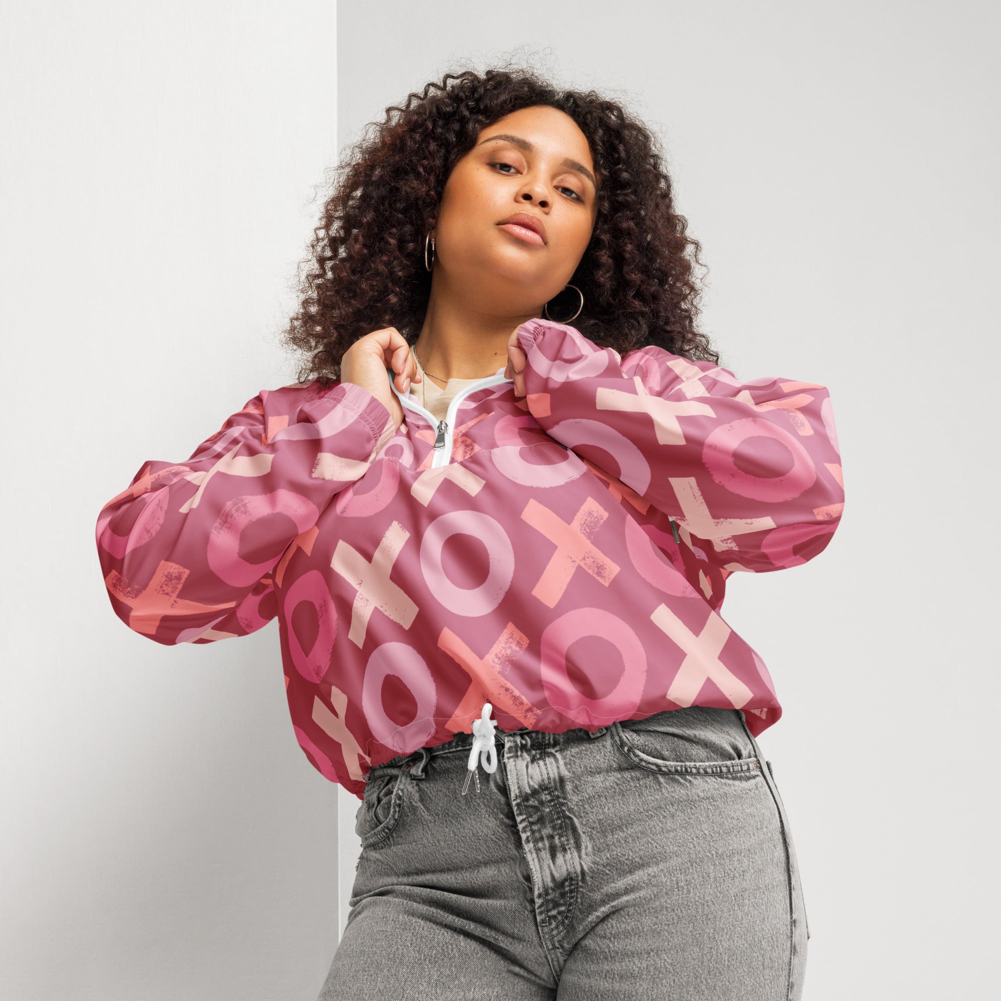 Ein Damen Cropped Windbreaker in verschiedenen Rosa- und Rottönen mit einem durchgängigen Muster aus großen X- und O-Formen. Die Jacke hat eine Kapuze, einen halben Reißverschluss, weiße Kordeln und einen verstellbaren Saum. Die Jacke ist flach ausgebreitet. Präsentiert von einer kräftigeren Frau in der Frontansicht by Just a Girl.
