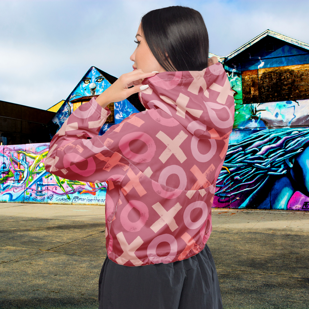 Ein Damen Cropped Windbreaker in verschiedenen Rosa- und Rottönen mit einem durchgängigen Muster aus großen X- und O-Formen. Die Jacke hat eine Kapuze, einen halben Reißverschluss, weiße Kordeln und einen verstellbaren Saum. Die Jacke ist flach ausgebreitet. Präsentiert von einer Frau in der Rückansicht by Just a Girl.