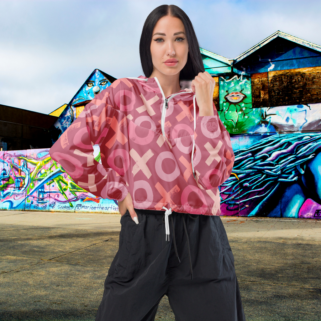 Ein Damen Cropped Windbreaker in verschiedenen Rosa- und Rottönen mit einem durchgängigen Muster aus großen X- und O-Formen. Die Jacke hat eine Kapuze, einen halben Reißverschluss, weiße Kordeln und einen verstellbaren Saum. Die Jacke ist flach ausgebreitet. Präsentiert von einer Frau in der Frontansicht by Just a Girl.