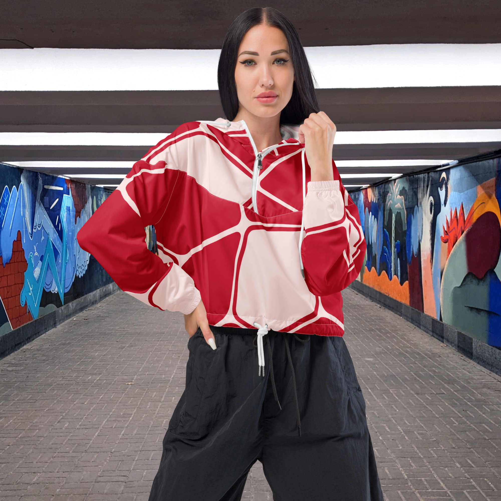 Ein Damen Cropped Windbreaker in Rot und hellem Rosé mit einem abstrakten geometrischen Muster aus miteinander verbundenen, abgerundeten Rechtecken und Linien. Die Jacke hat eine Kapuze, einen halben Reißverschluss, weiße Kordeln und einen verstellbaren Saum. Mockup Girl Vorder Ansicht. By Just a Girl.