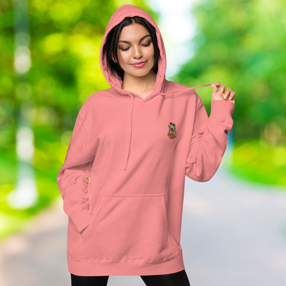 Ein weibliches Model zeigt lässig tragend den pigment pinken Kapuzenpullover mit der Vorderansicht im Freien. Die rechte Hand ist in der Kängurutasche. Auf der linken Brust befindet sich ein Frosch der auf einer Discokugel sitzt eine Ukulele hält und eine Herz  Sonnenbrille auf hat als Stickerei-Emblem, Dazu gibts noch die passende Sweatpants.  Designed, by Just a Girl.