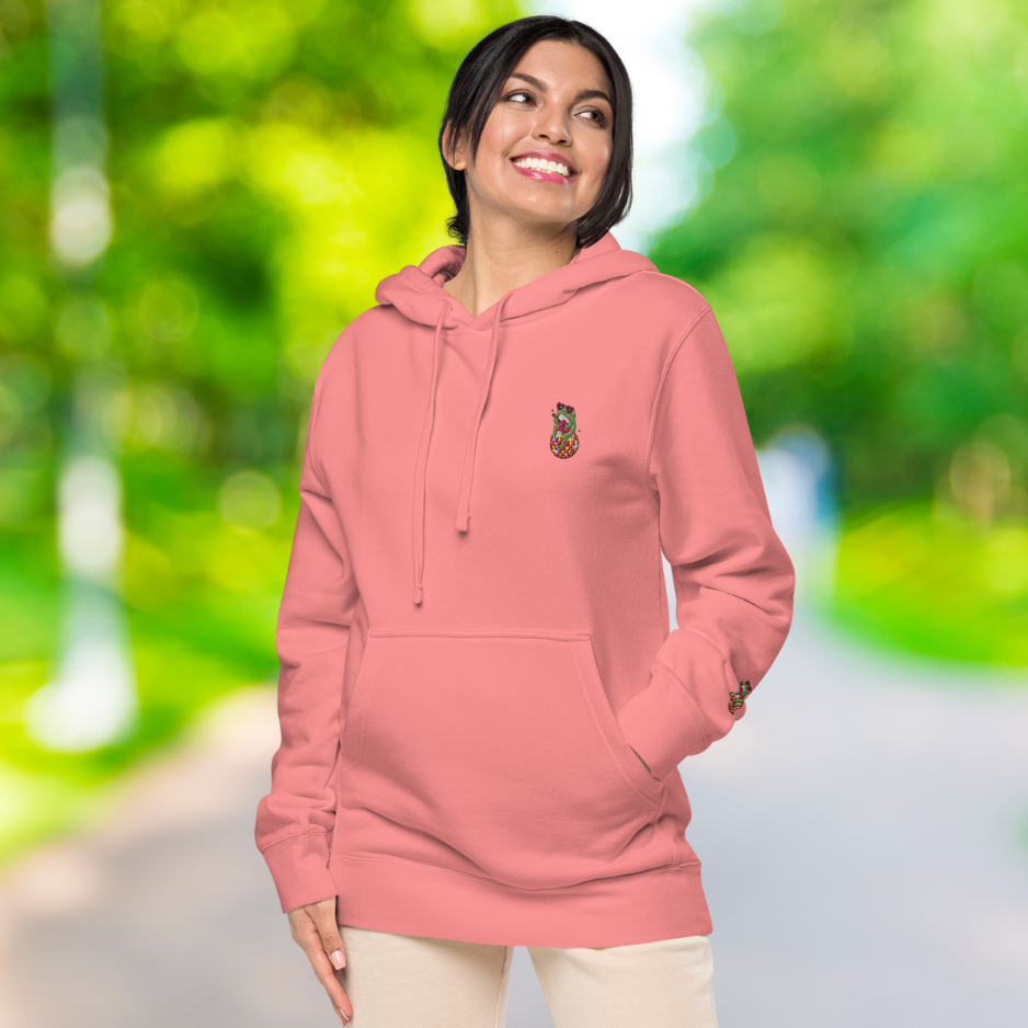 Ein weibliches Model zeigt lässig tragend den "Happy Frog" Kapuzenpullover seitliche linke Vorderansicht mit der linken Hand in der vorderen Känguruhtasche.