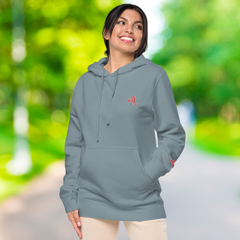 Ein weibliches Model zeigt sich draußen lässig von der linken Seite tragend den Kapuzenpullover in Schiefer blau "New York" mit Sicht auf das gestickte New Yorker Emblem auf der linken Brusthöhe.