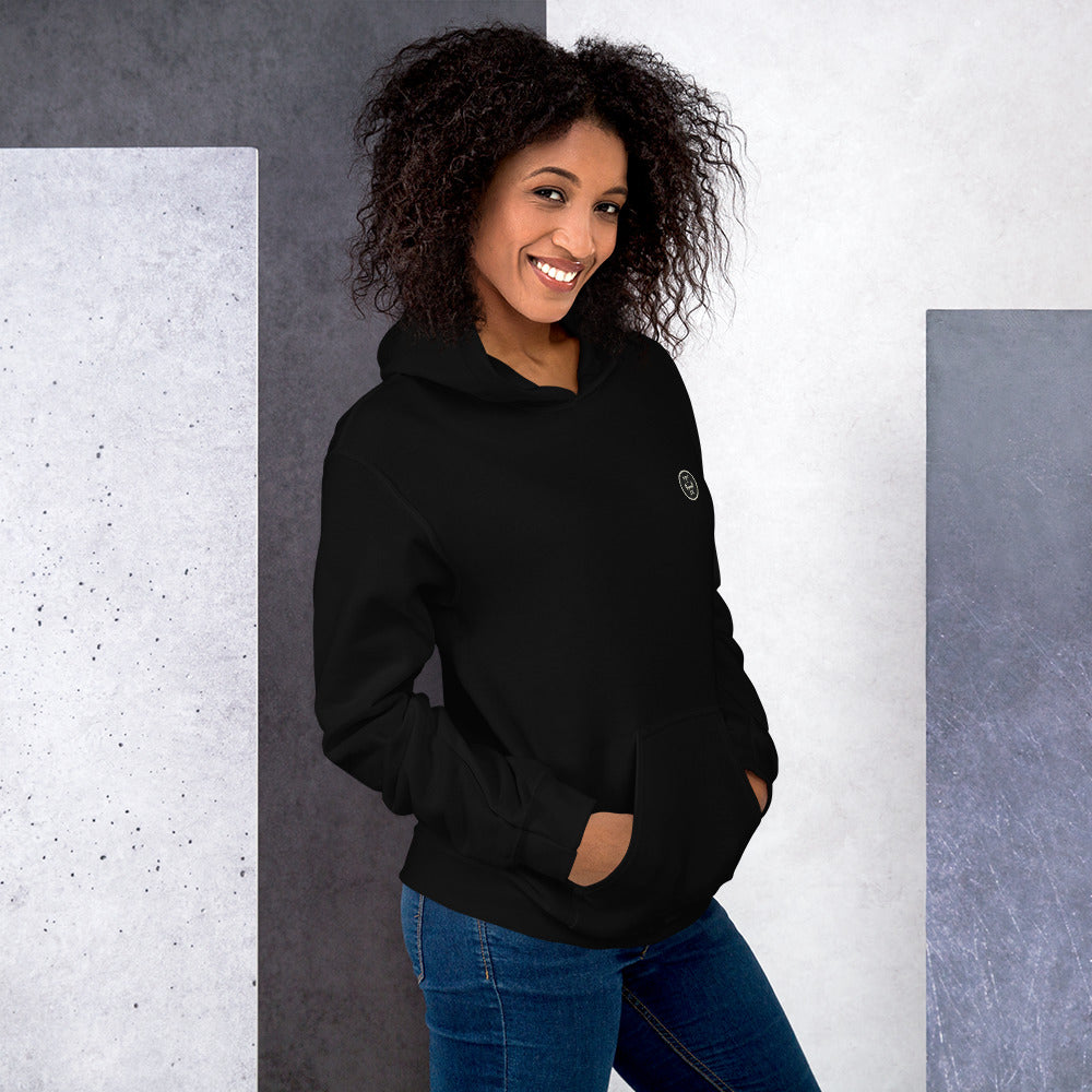 Ein weibliches Model lehnt lässig an einer Wand und zeigt tragend den "The Black One" Kapuzenpullover seitliche Vorderansicht mit beiden Händen in der vorderen Beuteltasche.