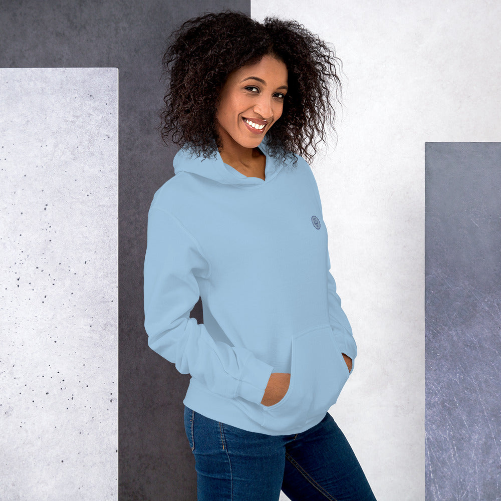 Ein weibliches Model lehnt lässig an einer Wand und zeigt tragend den "The Blue One" Kapuzenpullover seitliche Vorderansicht mit beiden Händen in der vorderen Beuteltasche.