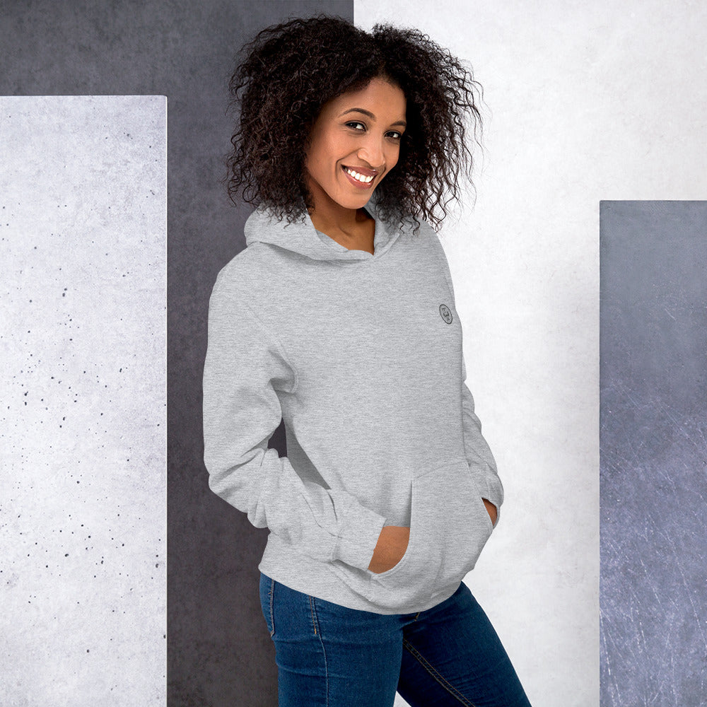 Ein weilbliches Model lehnt lässig an einer Wand und zeigt tragend den "The Grey One" Kapuzenpullover seitliche Vorderansicht mit beiden Händen in der vorderen Beuteltasche.