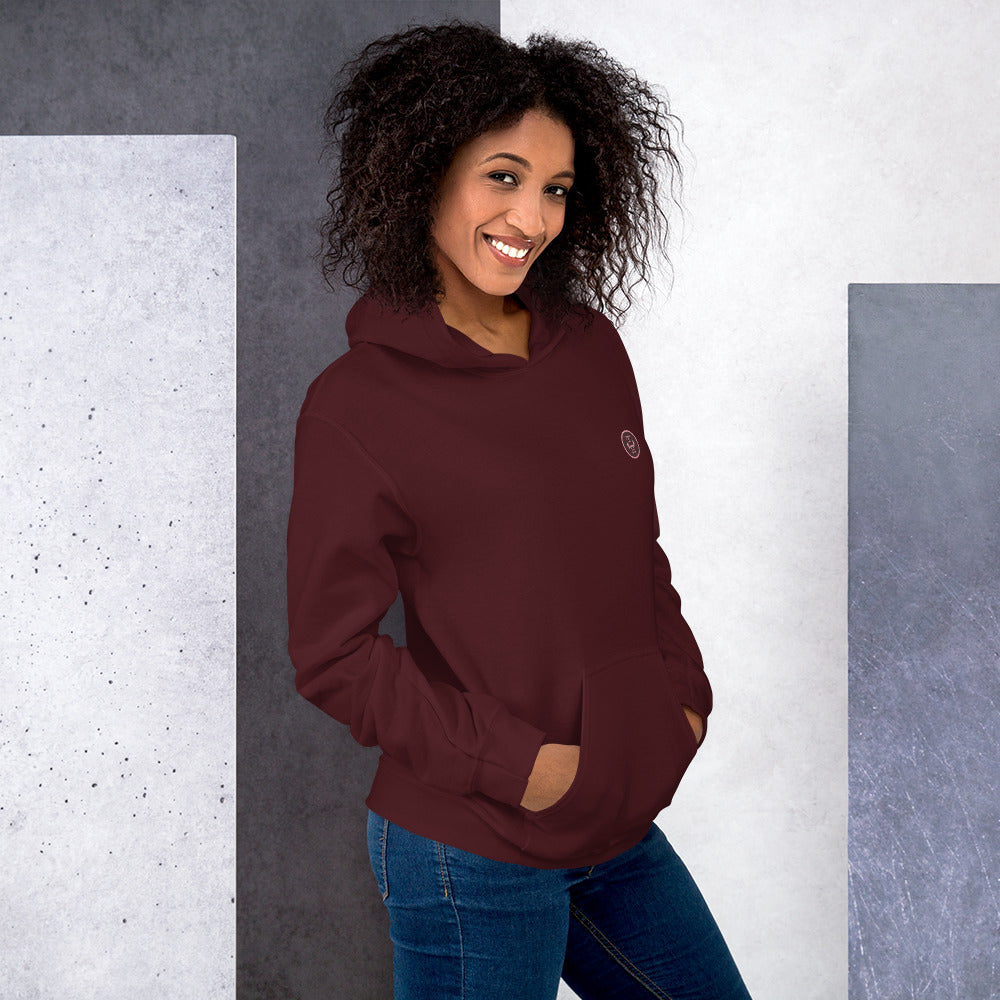 Ein weibliches Model lehnt lässig an einer Wand und zeigt tragend den "The Maroon One" Kapuzenpullover seitliche Vorderansicht mit beiden Händen in der vorderen Beuteltasche.