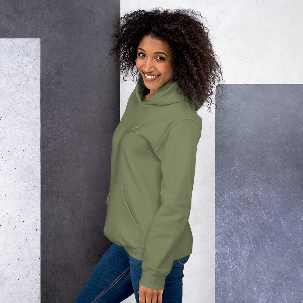 Ein weibliches Model zeigt sich lässig von der linken Seite tragend den Kapuzenpullover "The Olive One" mit Sicht auf das gestickte Eulen Emblem auf der linken Brusthöhe.