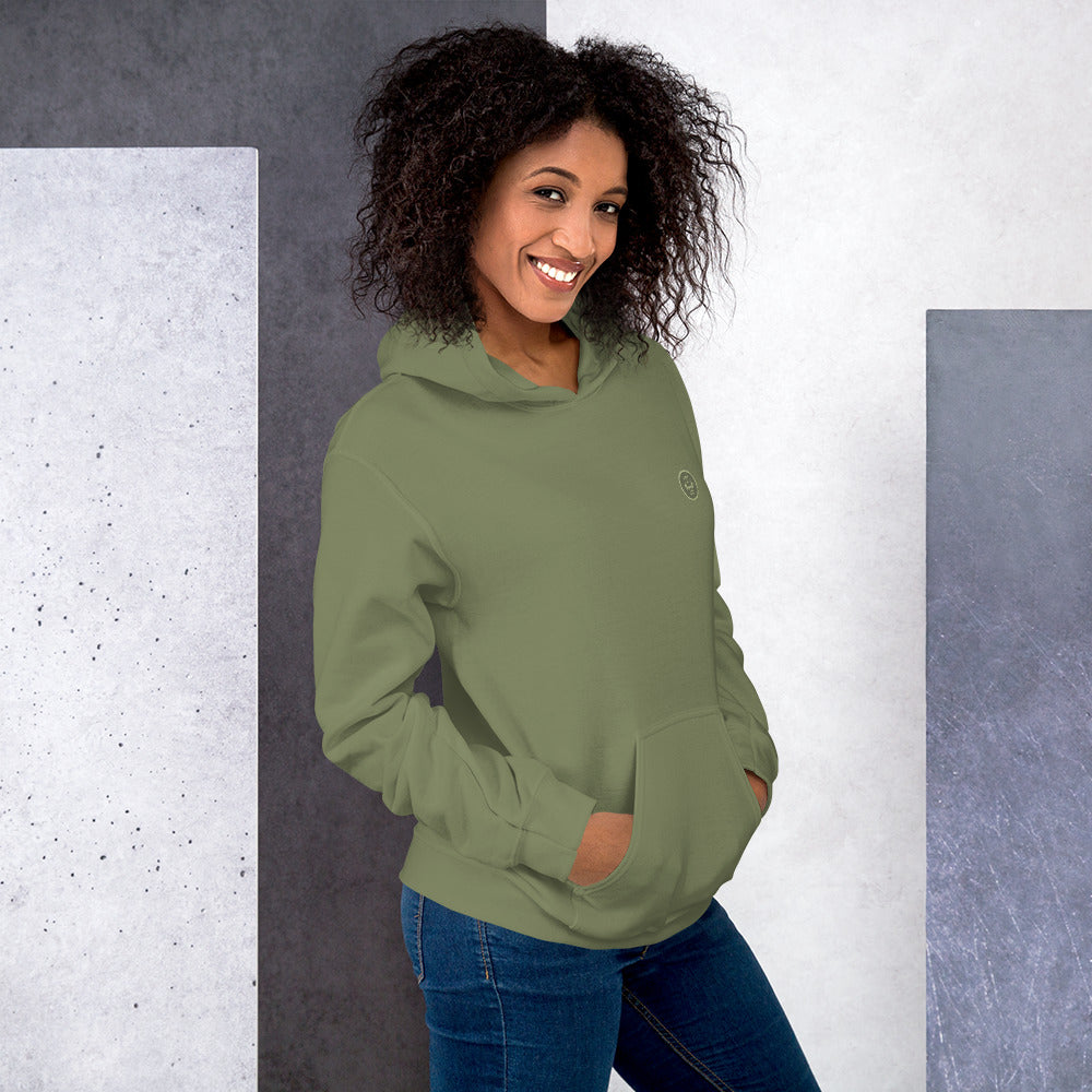 Ein weibliches Model lehnt lässig an einer Wand und zeigt tragend den "The Olive One" Kapuzenpullover seitliche Vorderansicht mit beiden Händen in der vorderen Beuteltasche.