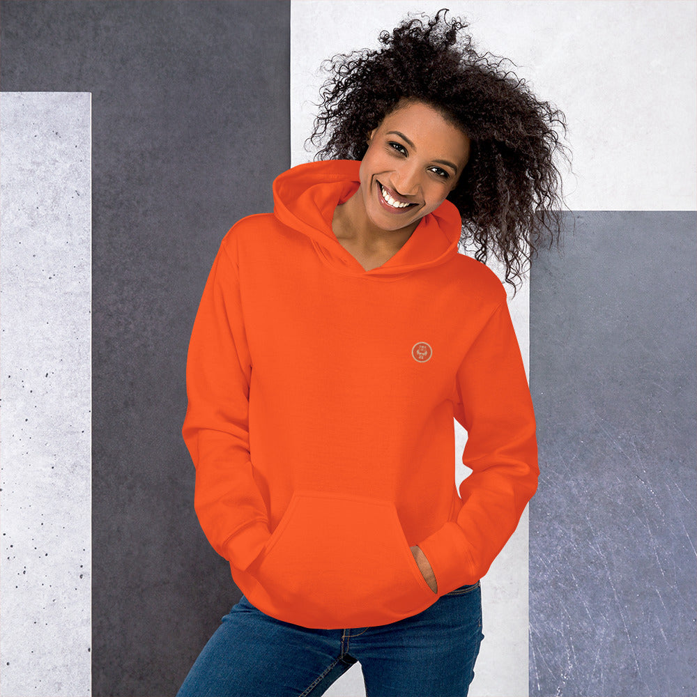 Ein weibliches Model zeigt sich mit dem Kapuzenpullover "The Orange One" Beide Hände lässig in der vorderen Beuteltasche.