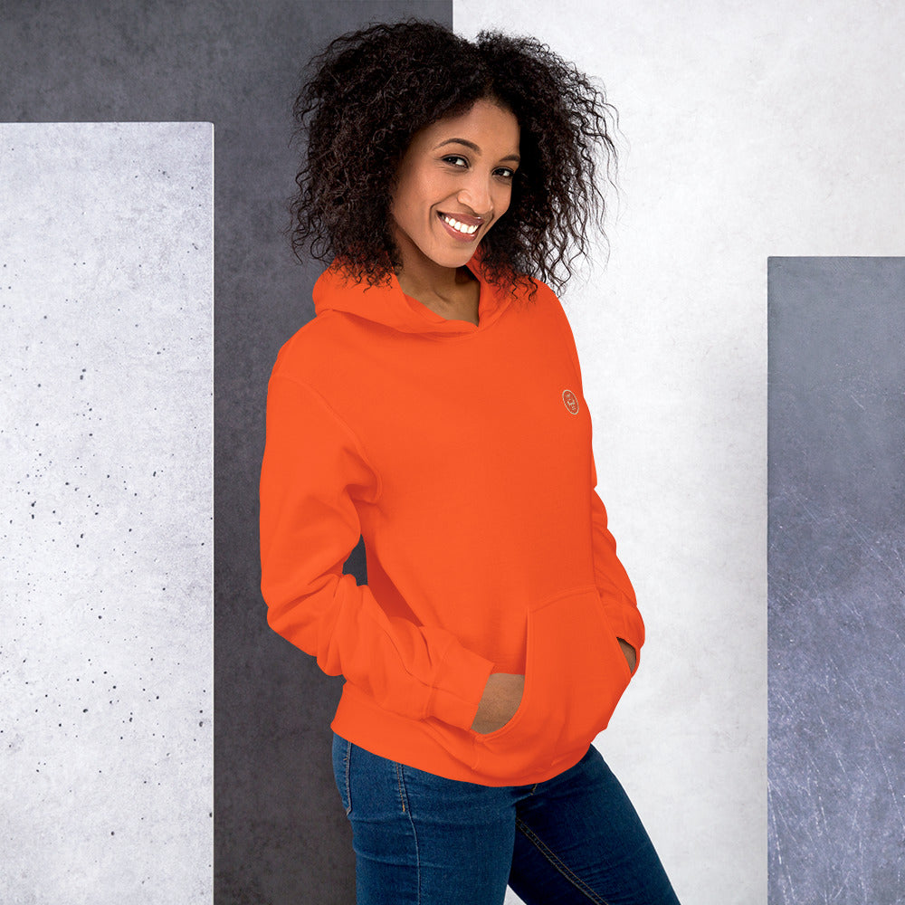 Ein weibliches Model lehnt lässig an einer Wand und zeigt tragend den "The Orange One" Kapuzenpullover seitliche Vorderansicht mit beiden Händen in der vorderen Beuteltasche.