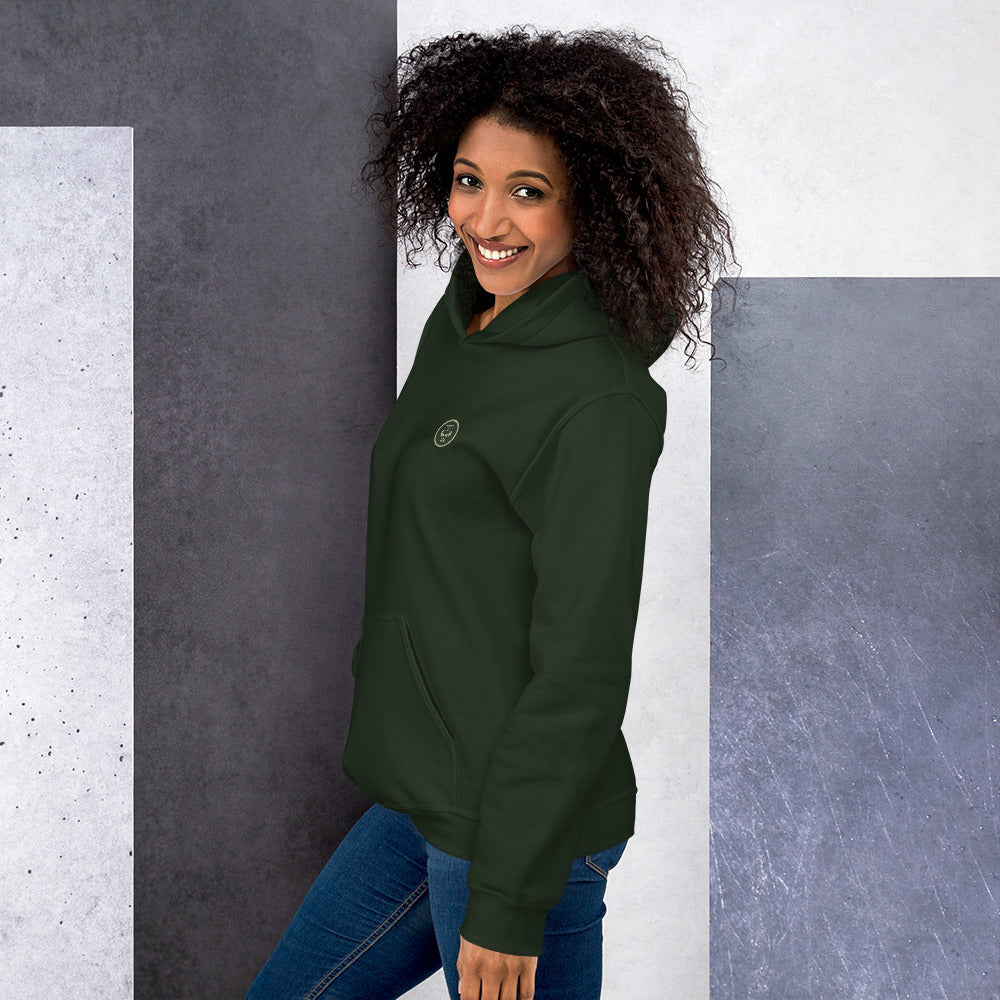 Ein weibliches Model zeigt sich lässig von der linken Seite tragend den Kapuzenpullover "The Pine One" mit Sicht auf das gestickte Eulen Emblem auf der linken Brusthöhe.