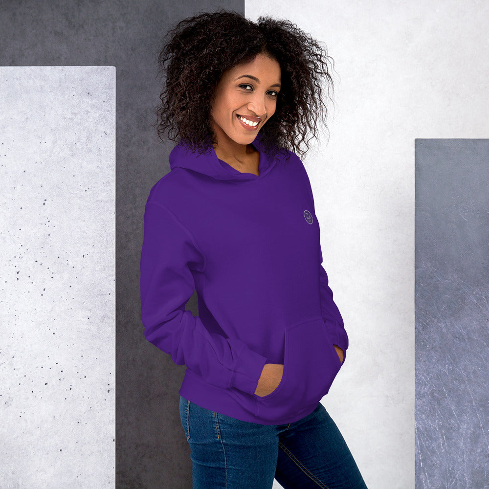 Ein weibliches Model lehnt lässig an einer Wand und zeigt tragend den "The Purple One" Kapuzenpullover seitliche Vorderansicht mit beiden Händen in der vorderen Beuteltasche.