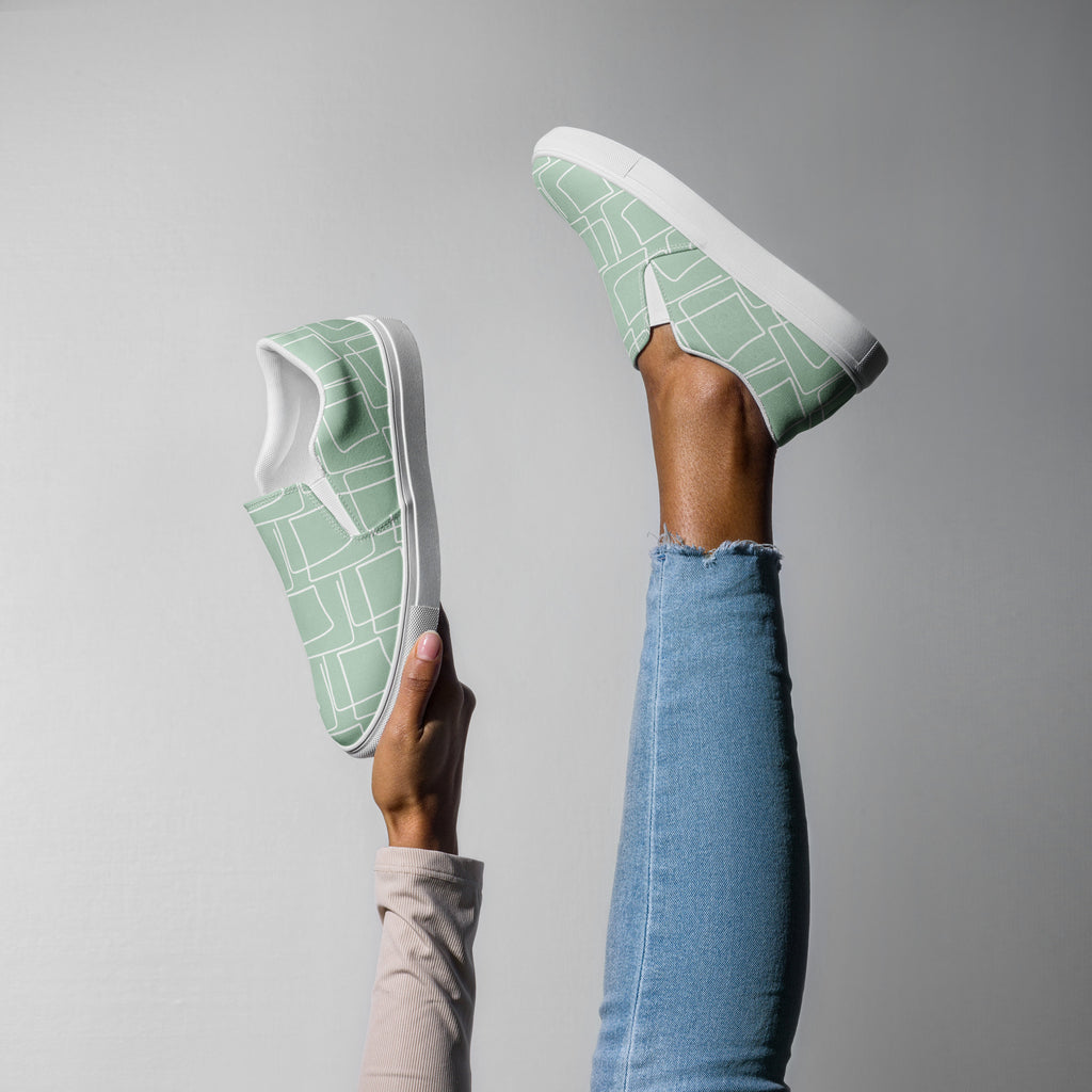 Ein weibliches Model trägt den Slip On Schuh Mint Maze streckt ein Bein in die Luft und hält in der anderen Hand den anderen Schuh, mit einem organischen, fließenden Muster in Minzgrünund weiß. Designed, by Just a Girl.