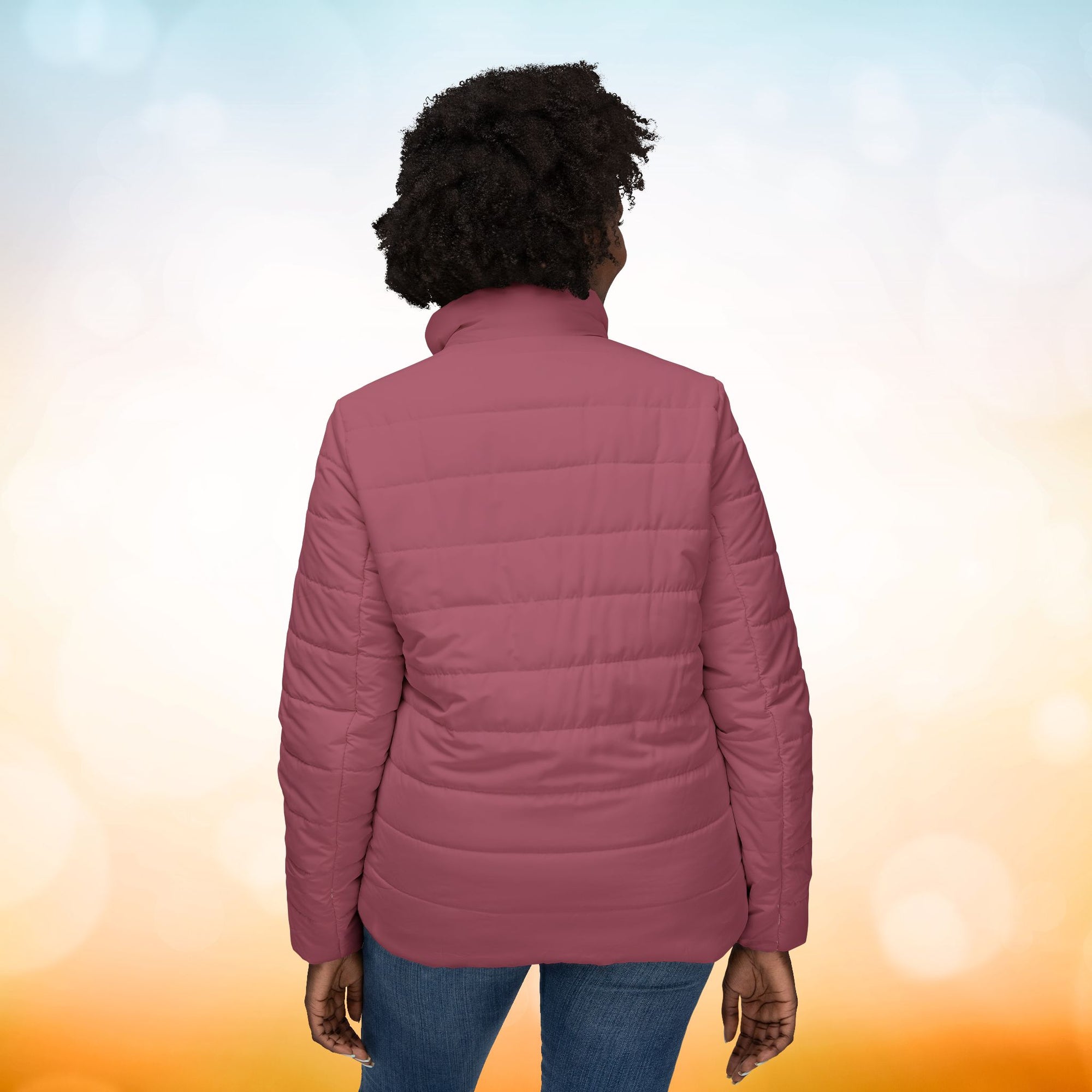 Ein weibliches Model zeigt sich von der Rückseite mit der  Damen-Pufferjacke in einem zarten Rosenstaub. Die Jacke ist horizontal gesteppt, hat eine glatte Oberfläche sowie einen hohen Stehkragen. Designed, by Just a Girl.