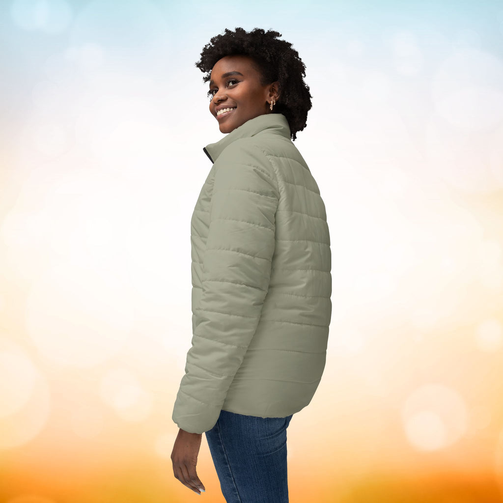  Ein weibliches Model trägt eine  Damen-Pufferjacke Ansicht von der Seite in einem Ash Grey Farbton. Die Jacke ist horizontal gesteppt, hat eine glatte Oberfläche, einen hohen Stehkragen und einen durchgehenden Reißverschluss. Designed, by Just a Girl