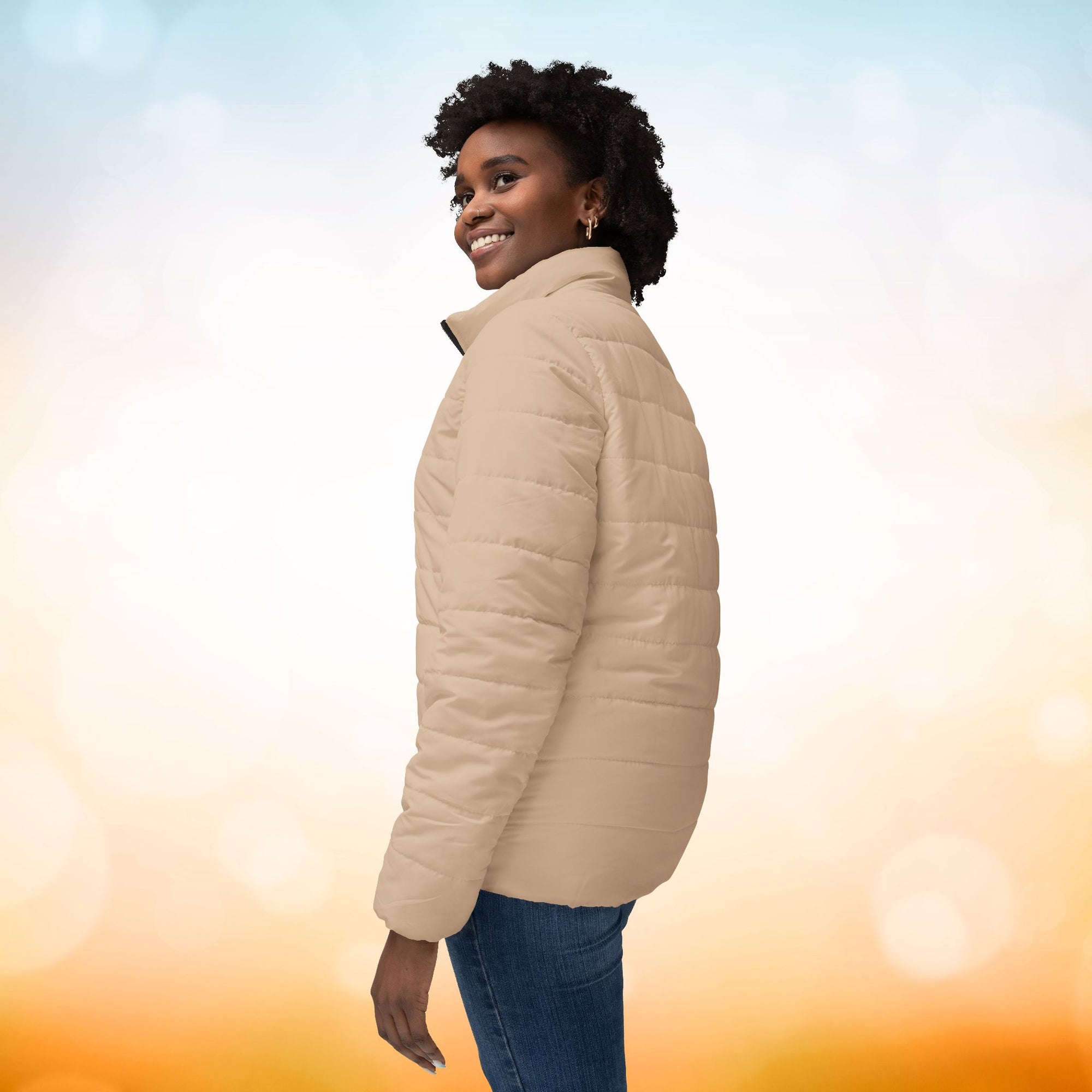 Ein weibliches Model zeigt die Damen-Pufferjacke von der linken Seite in einem Brown Sugar Farbton. Die Jacke ist horizontal gesteppt, hat eine glatte Oberfläche, einen hohen Stehkragen und einen durchgehenden Reißverschluss. designed, by Just a Girl.

