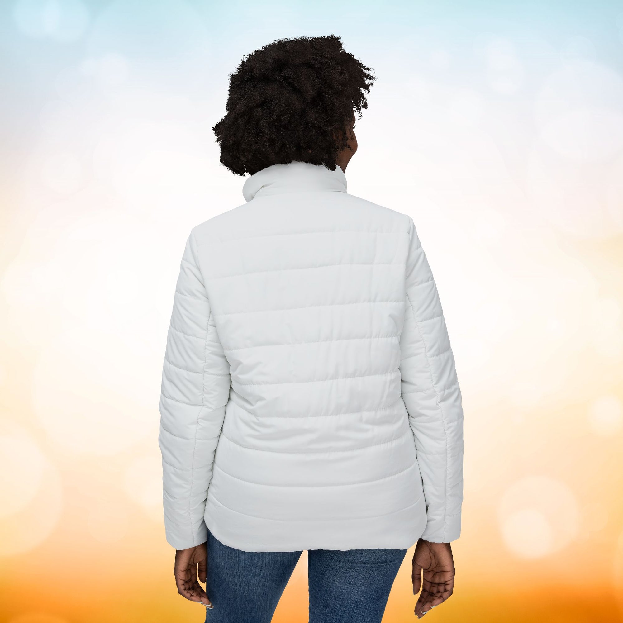 Ein weibliches Model zeigt sich von der Rückenansicht mit einer Damen-Pufferjacke  in einem Ivory Farbton. Die Jacke ist horizontal gesteppt, hat eine glatte Oberfläche, einen hohen Stehkragen und einen durchgehenden Reißverschluss. Designed, by Just a Girl

