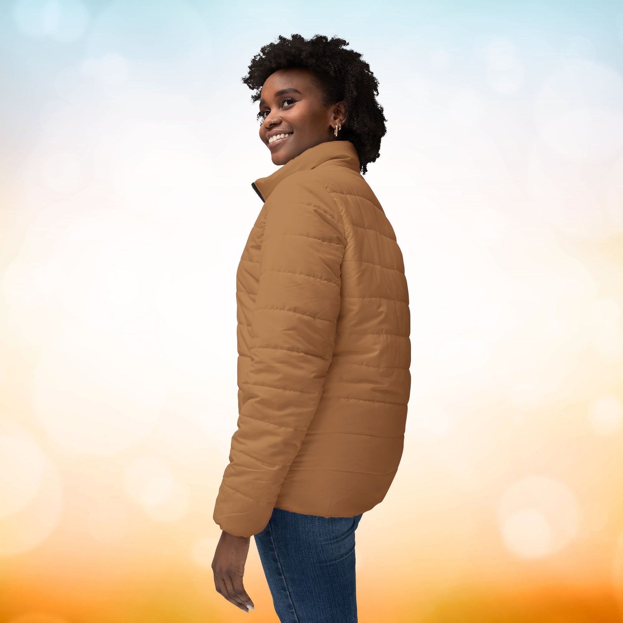 Ein weibliches Model zeigt die Damen-Pufferjacke mit der linken Seitenansicht in einem Persian-Orange farbton. Die Jacke ist horizontal gesteppt, hat eine glatte Oberfläche, einen hohen Stehkragen und einen durchgehenden Reißverschluss. Designed, by Just a Girl.

