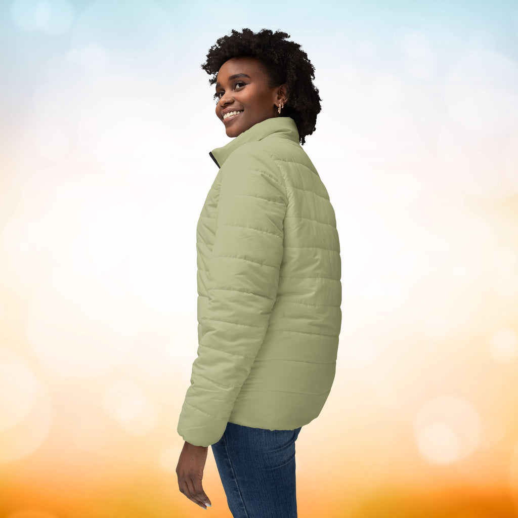 Ein weibliches Model trägt eine Damen-Pufferjacke in Sage Green. Das Model zeigt sich von der linken Seitenansicht.  Die Jacke ist horizontal gesteppt, hat eine glatte Oberfläche, einen hohen Stehkragen und einen durchgehenden Reißverschluss. Designed, by Just a Girl.