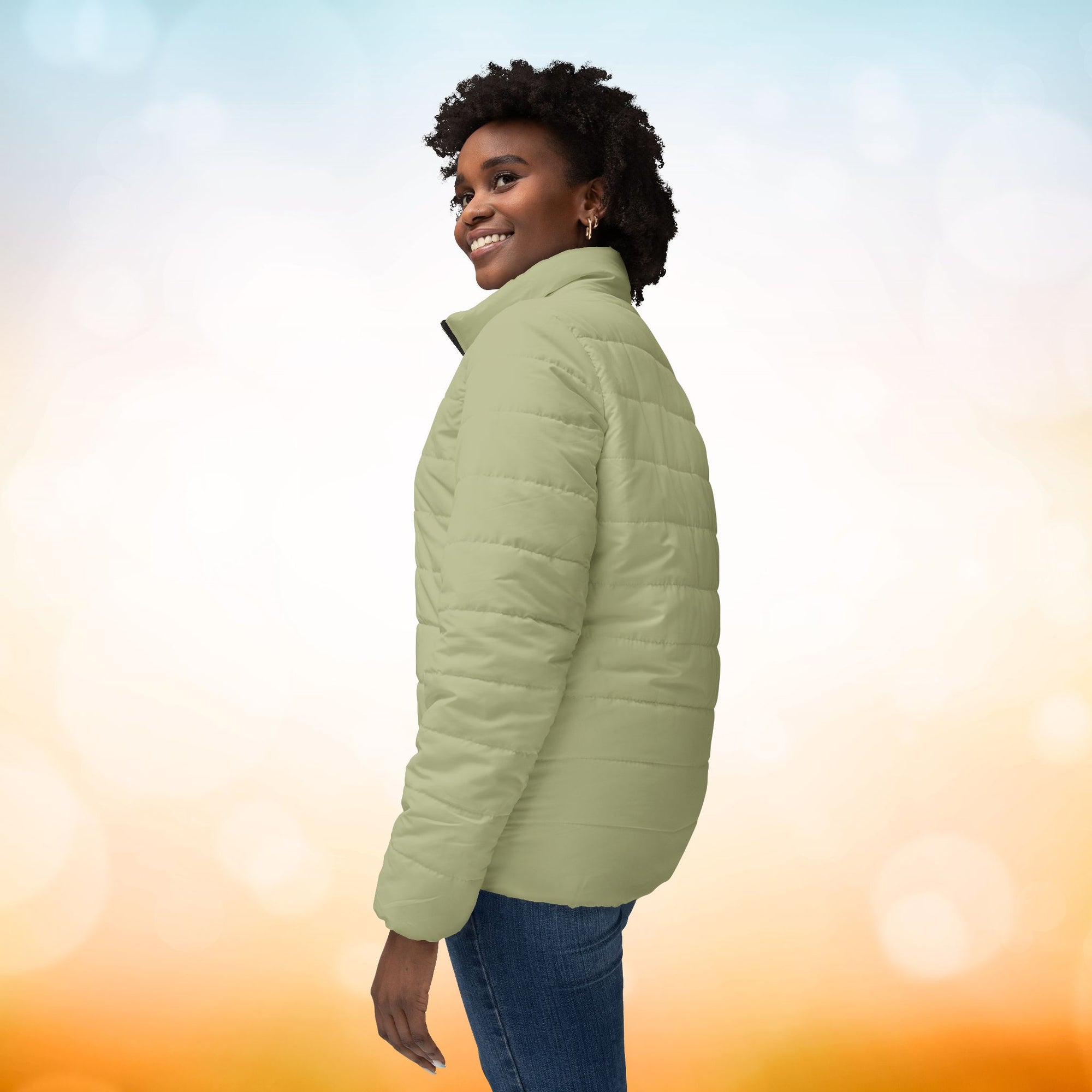 Ein weibliches Model trägt eine Damen-Pufferjacke in Sage Green. Das Model zeigt sich von der linken Seitenansicht.  Die Jacke ist horizontal gesteppt, hat eine glatte Oberfläche, einen hohen Stehkragen und einen durchgehenden Reißverschluss. Designed, by Just a Girl.