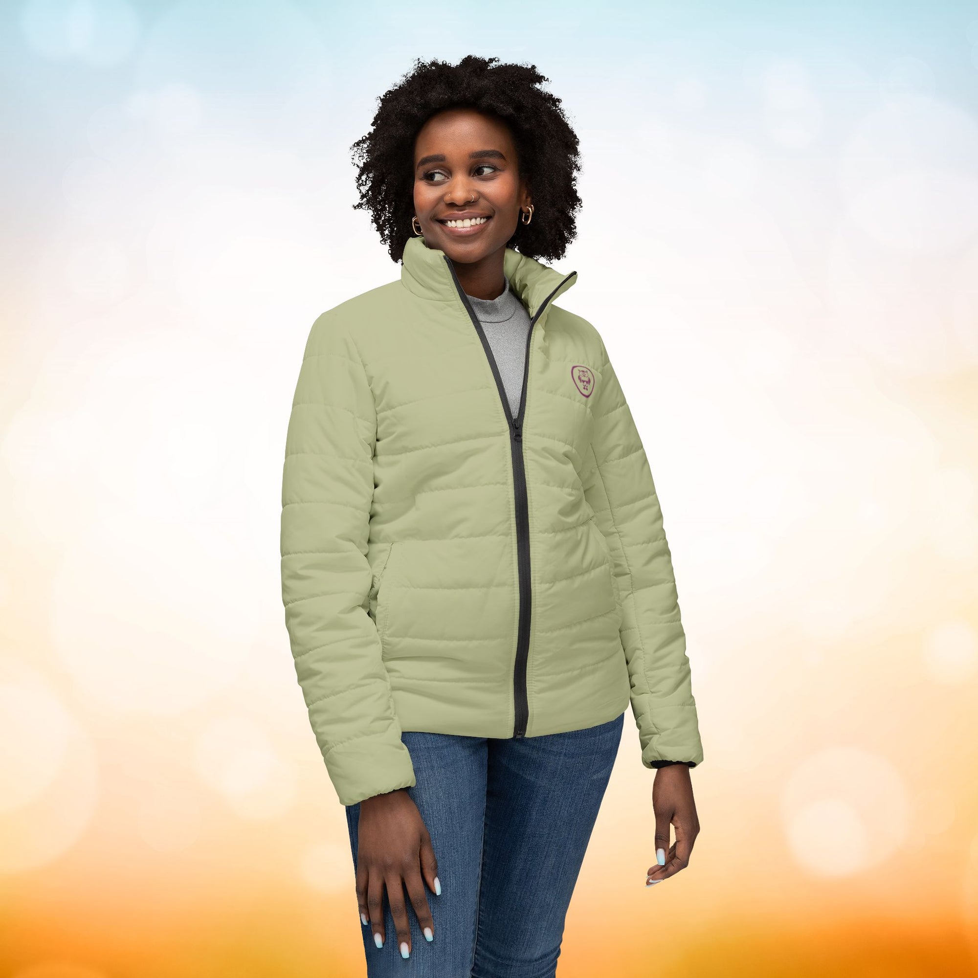 Ein weibliches Model trägt eine Damen-Pufferjacke in Sage Green. Das Model zeigt sich von der Vorderansicht. Die Jacke ist horizontal gesteppt, hat eine glatte Oberfläche, einen hohen Stehkragen und einen durchgehenden Reißverschluss. Designed, by Just a Girl.