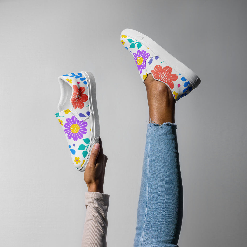 Ein weibliches Model zeigt tragend mit einem Bein in die Luft gestreckt und mit der anderen Hand halten die Slip-on-Schuhe mit einem Muster aus bunten, stilisierten Blumen in Rot, Lila, Gelb, Grün und Blau.  Designed, by Just a Girl.