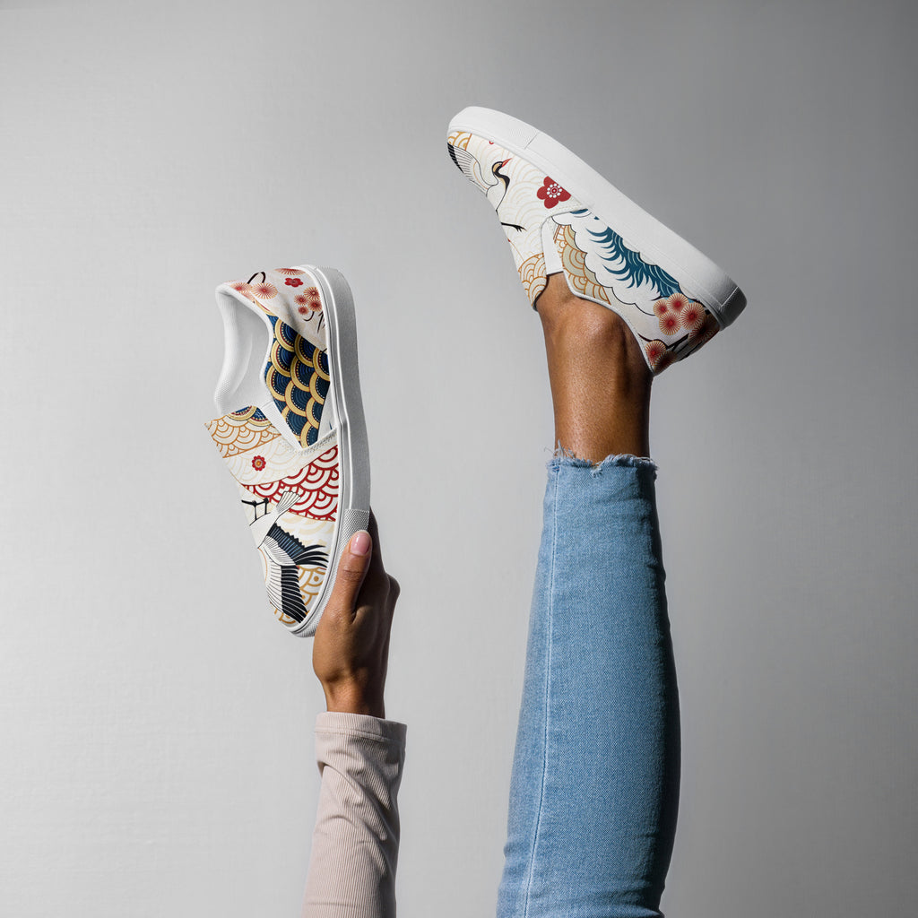 Ein weibliches Model zeigt tragend ein Bein in die Luft gestreckt und mit der anderen Hand den 2. Schuh zeigend die  Damen-Slip-on-Schuhe mit einem bunten Muster im japanischen Stil. Das Motiv zeigt Kraniche, Wellen, Kirschblüten und geometrische Muster. Designed, by Just a Girl