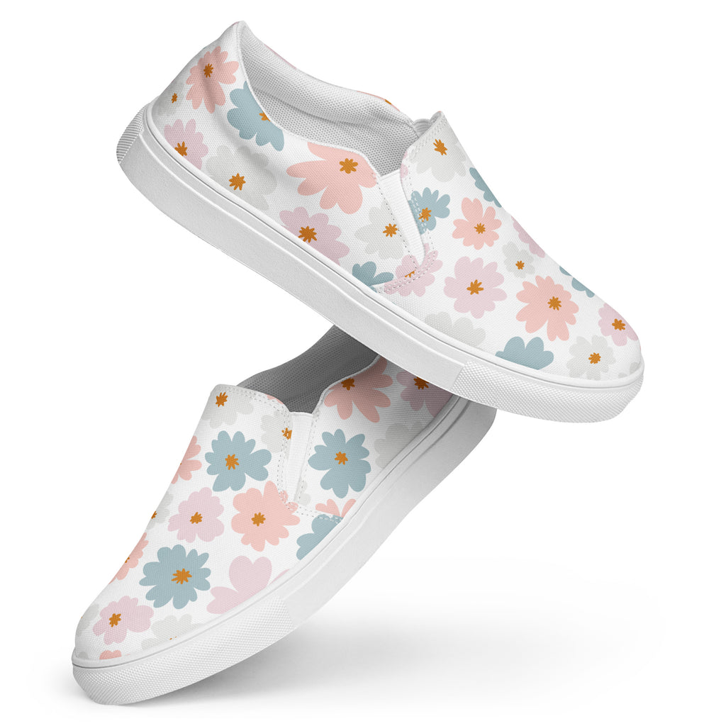 Ein Paar weiße Damen-Slip-on-Schuhe mit einem Muster aus pastellfarbenen Blüten in Hellblau und Zartrosa. Die Schuhe schweben vor einem weißen Hintergrund. Designed, by Just a Girl.