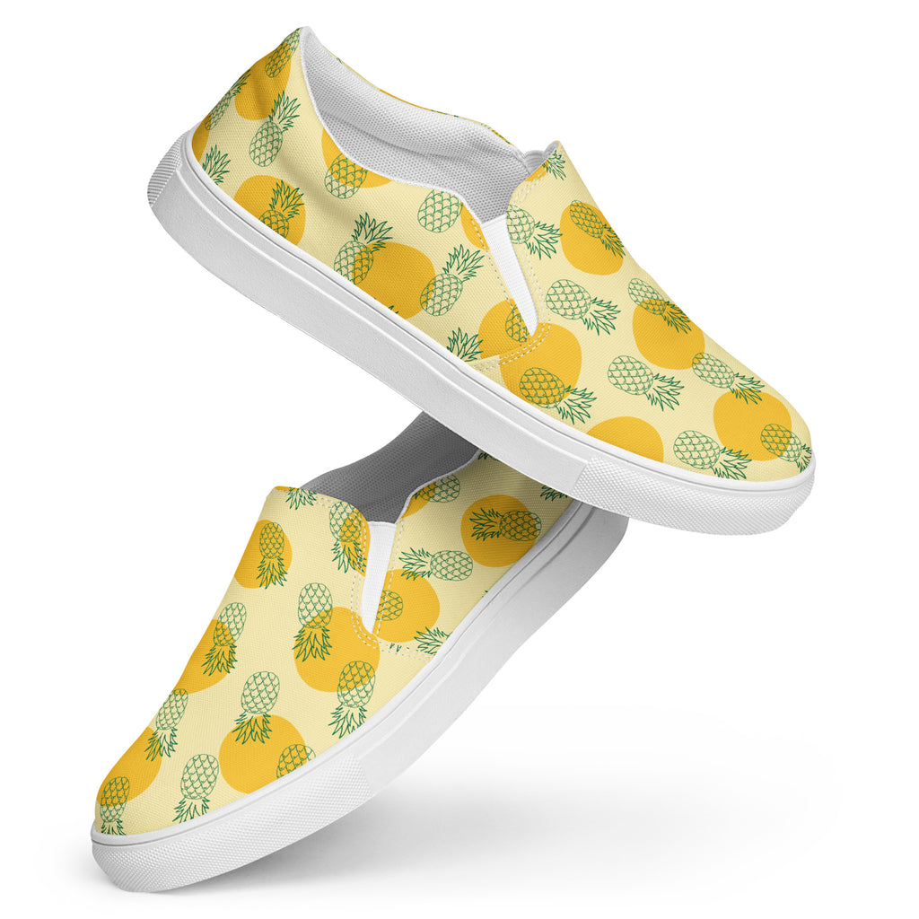 Ein Paar Damen Slip-on-Schuhe mit einem Muster aus grünen Ananas-Outlines und orangen Kreisen auf gelben Hintergrund. Die Schuhe schweben vor einem weißen Hintergrund. Designed, by Just a Girl.