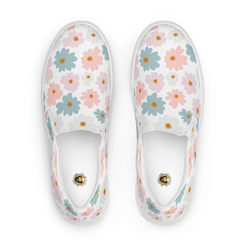 Ein Paar weiße Damen-Slip-on-Schuhe mit einem Muster aus pastellfarbenen Blüten in Hellblau und Zartrosa. Die Schuhe werden von der Ansicht von oben  vor einem weißen Hintergrund gezeigt. Designed, by Just a Girl.