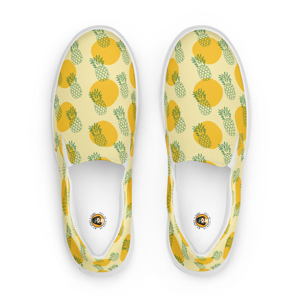 Ein Paar Damen Slip-on-Schuhe mit einem Muster aus grünen Ananas-Outlines und orangen Kreisen auf gelben Hintergrund. Die Ansicht der Schuhe ist von oben vor einem weißen Hintergrund. Designed, by Just a Gir
