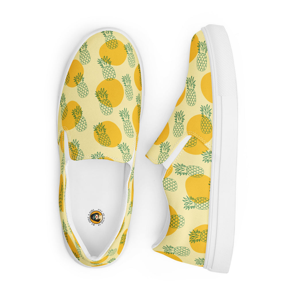 Ein Paar Damen Slip-on-Schuhe mit einem Muster aus grünen Ananas-Outlines und orangen Kreisen auf gelben Hintergrund. Die Ansicht der Schuhe ist ein Schuh von oben der andere liegt seitliche daneben vor einem weißen Hintergrund. Designed, by Just a Girl.