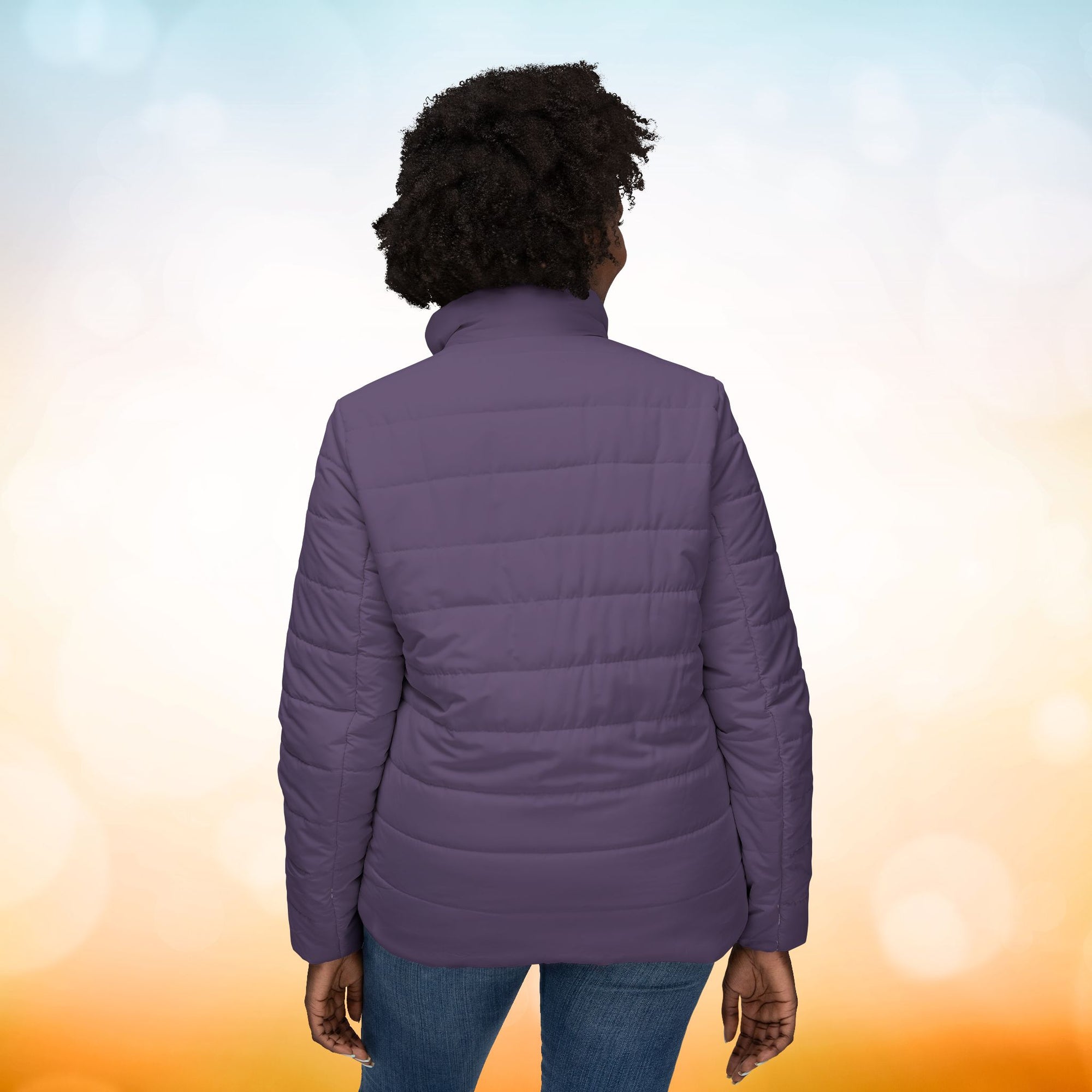 Das weibliche Model trägt eine  Damen-Pufferjacke in einem dunklem Dusty Plum Farbton. Die Jacke ist horizontal gesteppt, hat eine glatte Oberfläche, einen hohen Stehkragen sie präsentiert die Jacke von der Rückenansicht. Designed, by Just a Girl.