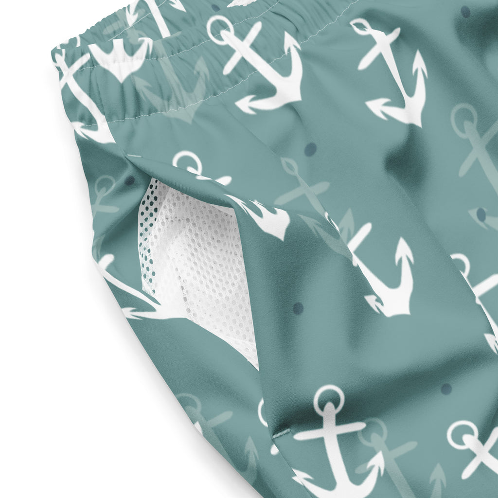 Eine Badeshorts in einem gedeckten Türkis- oder Blauton mit einem wiederkehrenden Muster aus weißen und hellgrauen Ankern und kleinen blauen Punkten. Der Bund ist elastisch und farblich passend.