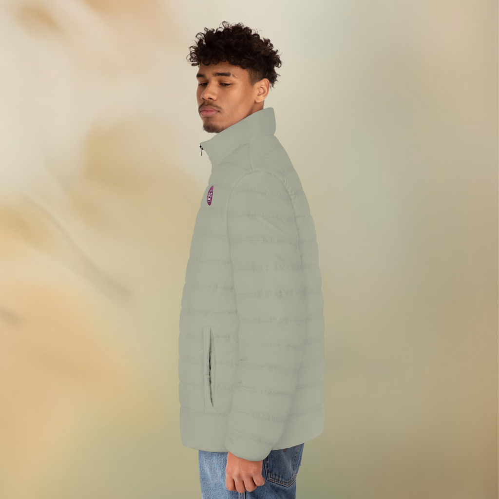 Ein männliches Model zeigt tragend von der linken Seite die Herren-Pufferjacke in einem Aschgrauen Farbton. Die Jacke ist horizontal gesteppt, hat eine glatte Oberfläche, einen hohen Stehkragen und einen durchgehenden Reißverschluss der Schwarz ist . Auf der linken Brusthöhe ist ein Emblem im Maori Stil. Designed, by Just a Girl