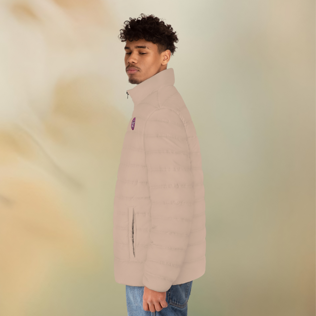 Ein männliches Model zeigt tragend mit der linken Seitenansicht die  Herren-Pufferjacke in einem Wüstensand Farbton. Die Jacke ist horizontal gesteppt, hat eine glatte Oberfläche, einen hohen Stehkragen und einen durchgehenden Reißverschluss der Schwarz ist . Auf der linken Brusthöhe ist ein Emblem im Maori Stil. Designed, by Just a Girl

